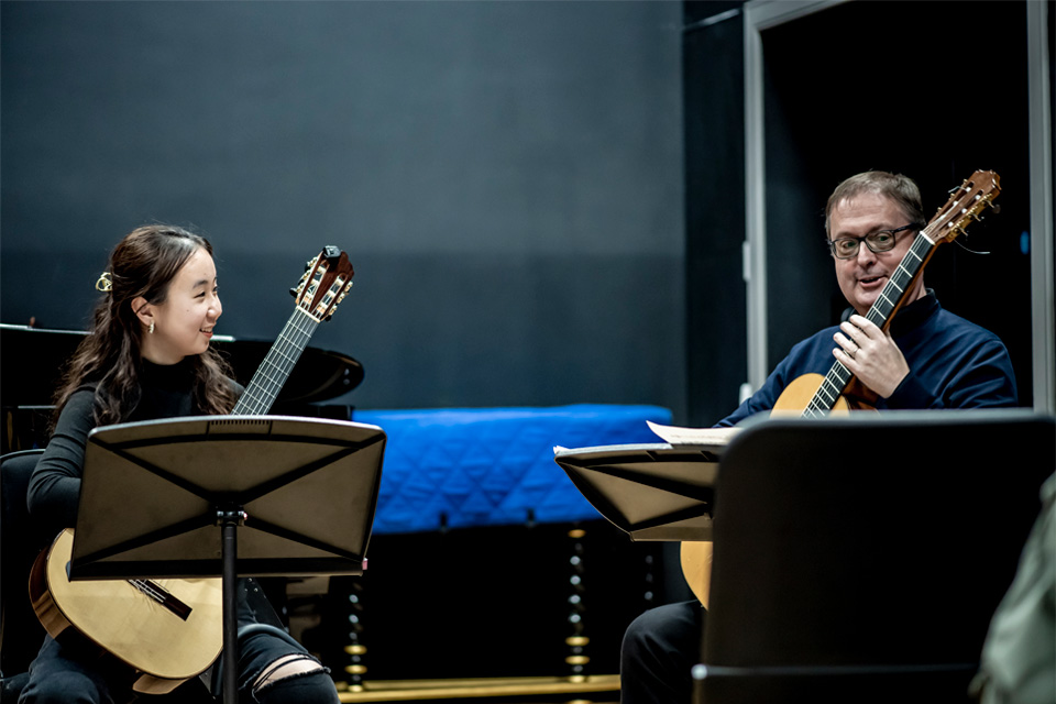 A man wearing a smart suit, holding a guitar, alongside a female Asian student, wearing a black top, also holding a guitar.